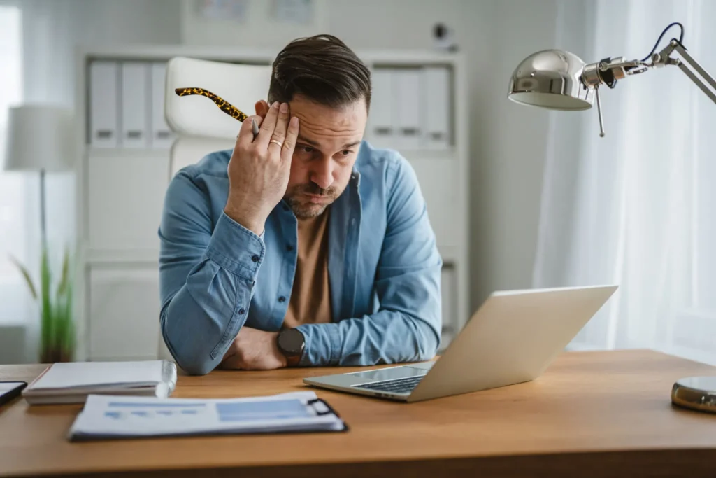 Stressed employee sitting at a home desk looking anxiously at a laptop screen
